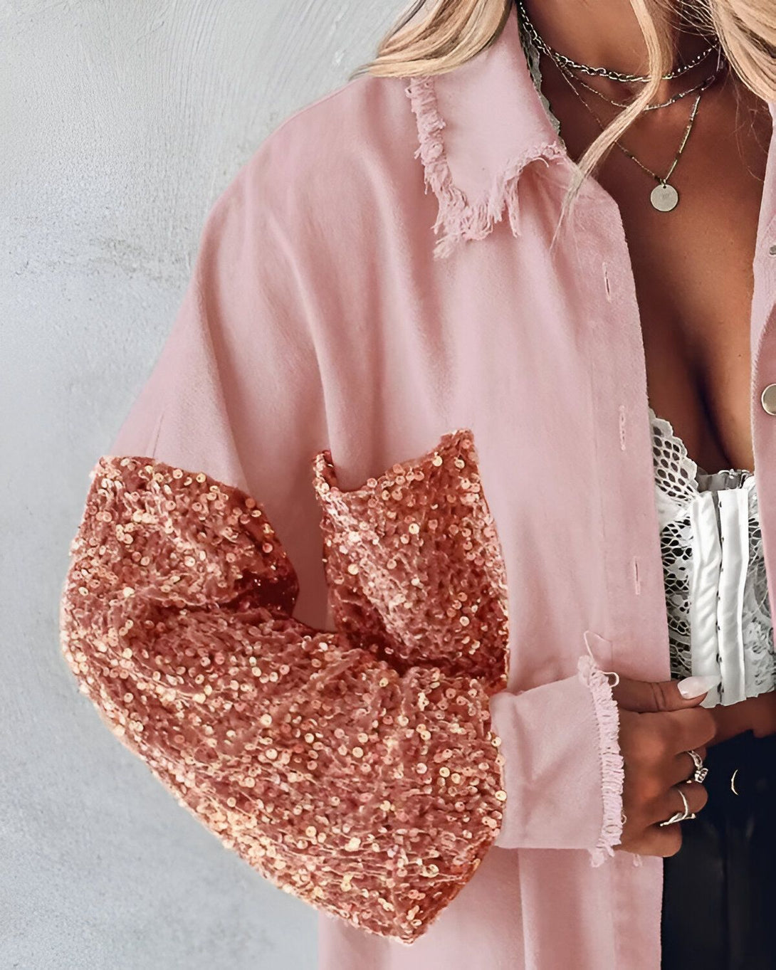 Une femme porte une chemise rose à manches longues. Les manches sont recouvertes de sequins dorés. La chemise est ouverte et on peut voir un soutien-gorge en dentelle blanc en dessous. La femme porte également un collier avec un pendentif rond.