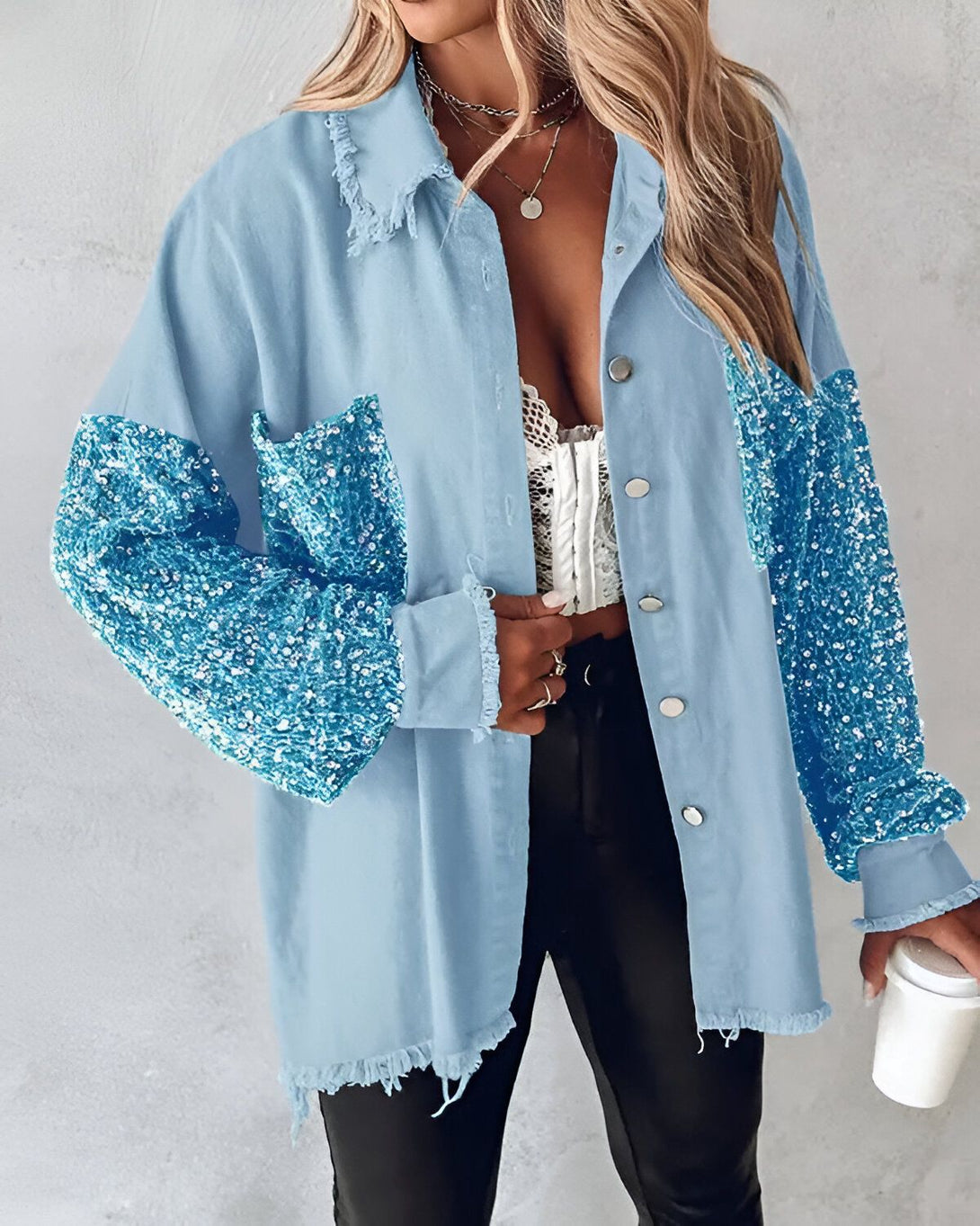 Une jeune femme porte une chemise en jean bleu clair avec des manches à sequins bleus. Elle est ouverte sur un haut blanc en dentelle. Elle porte également un pantalon noir et des talons noirs. Elle a les cheveux longs et blonds et porte un collier. Elle tient une tasse de café à la main.