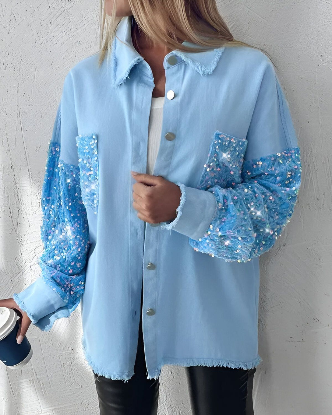 Une jeune femme porte une chemise en jean bleu clair avec des manches à sequins bleus. Elle est debout, de face, et tient une tasse de café à la main droite.