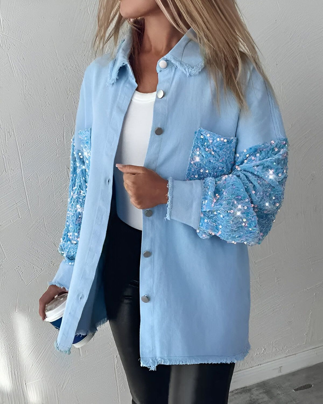 Une femme porte une veste en jean bleu clair avec des manches à sequins bleus. Elle porte également un pantalon en cuir noir et un haut blanc. Elle a les cheveux blonds et porte un maquillage léger. Elle tient une tasse de café à la main.