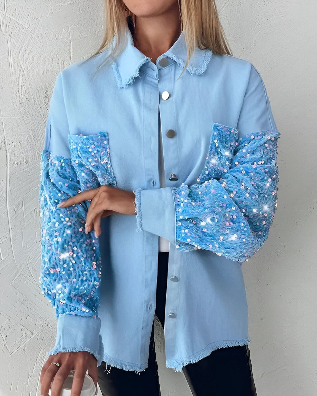 Une jeune femme porte une chemise en jean bleu clair avec des manches bouffantes en sequins bleus. Elle est ouverte et on peut voir un t-shirt noir en dessous. La chemise est courte et s'arrête au niveau de la taille. La femme a les cheveux blonds et elle est maquillée. Elle porte un collier et des boucles d'oreilles. Elle est debout et elle a une main dans la poche de sa chemise.