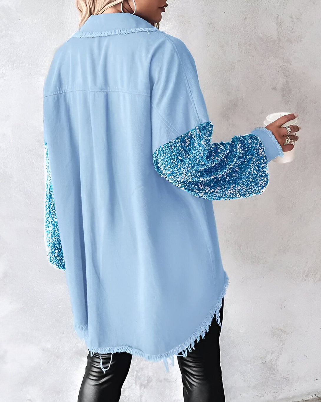Une femme porte une chemise en jean bleue avec des sequins sur les manches. La chemise est ouverte et on peut voir un t-shirt noir en dessous. Elle porte également un pantalon en cuir noir et des talons hauts noirs. Elle a les cheveux longs et noirs et porte un maquillage léger. Elle est debout dans un studio et tient une tasse de café à la main.