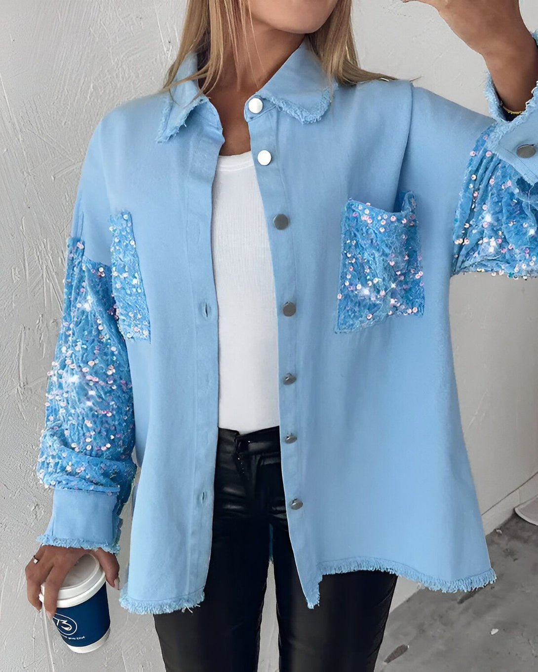 Une femme porte une chemise bleue en jean avec des sequins sur les manches et les poches. Elle porte également un pantalon en cuir noir et un t-shirt blanc. Elle tient une tasse de café à la main.