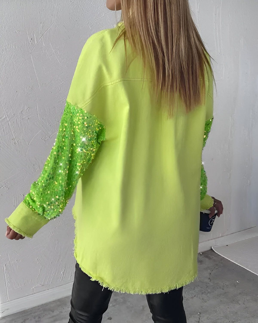 Une femme porte une chemise verte à manches longues. Les manches sont recouvertes de paillettes vertes. La chemise est ouverte et on peut voir un t-shirt noir en dessous. La femme est blonde et porte un pantalon noir. Elle est vue de dos.