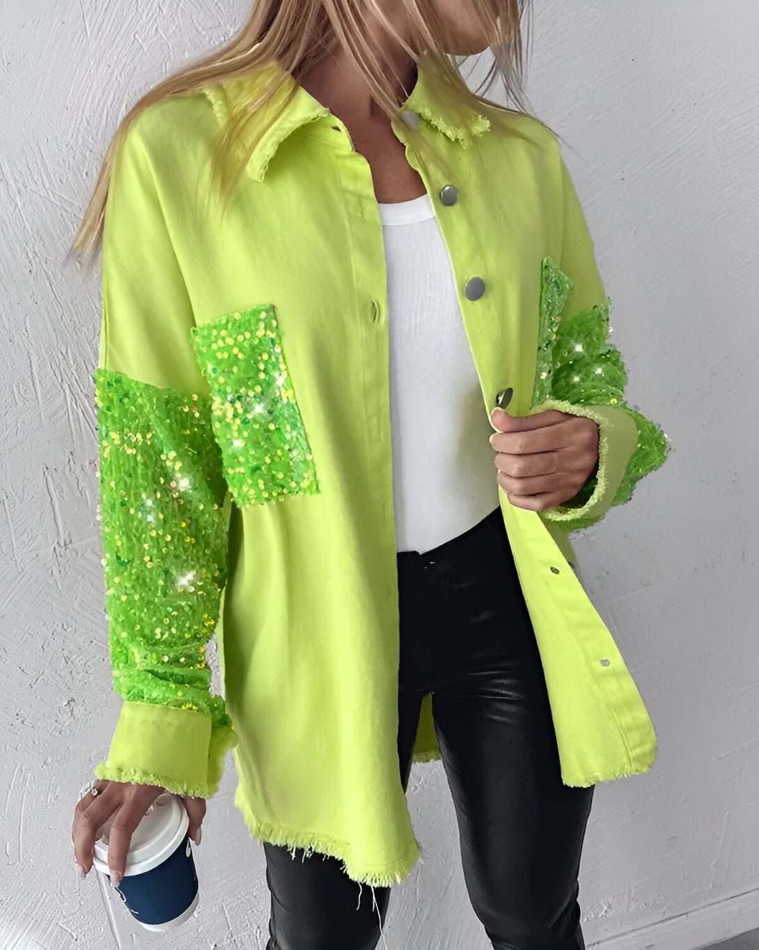 Une femme porte une chemise verte avec des paillettes sur les manches et un jean noir. Elle tient une tasse de café à la main.