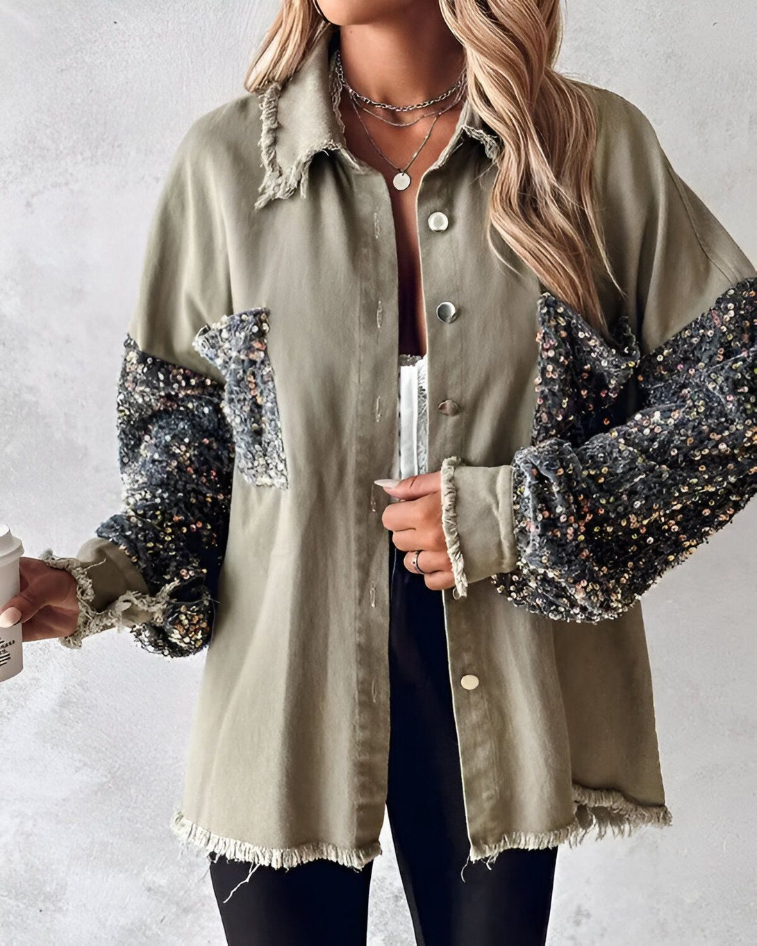 Une femme porte une veste verte kaki avec des sequins noirs et argentés sur les manches. La veste est ouverte et on peut voir un t-shirt blanc en dessous. Elle porte également un collier et un bracelet.