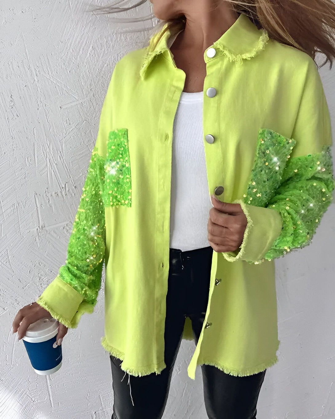 Une femme porte une veste en jean vert citron avec des sequins sur les manches et le devant. Elle porte également un pantalon noir et un haut blanc. Elle tient une tasse de café à la main.