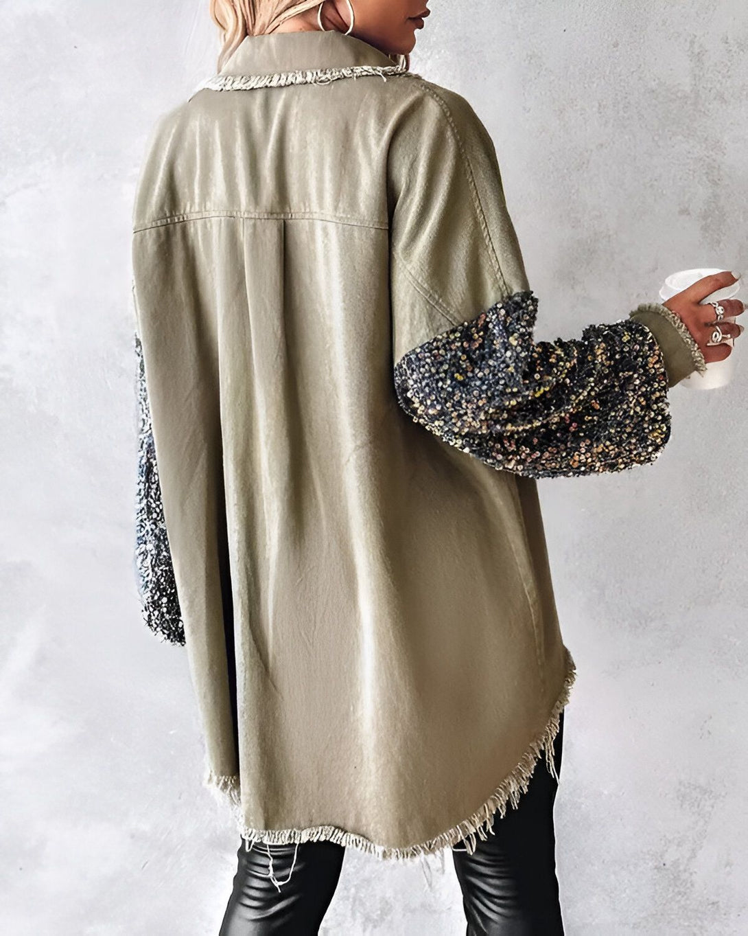 Une femme porte une chemise verte kaki avec des manches à sequins noirs et argentés. Elle est vue de dos et tient une tasse blanche à la main.
