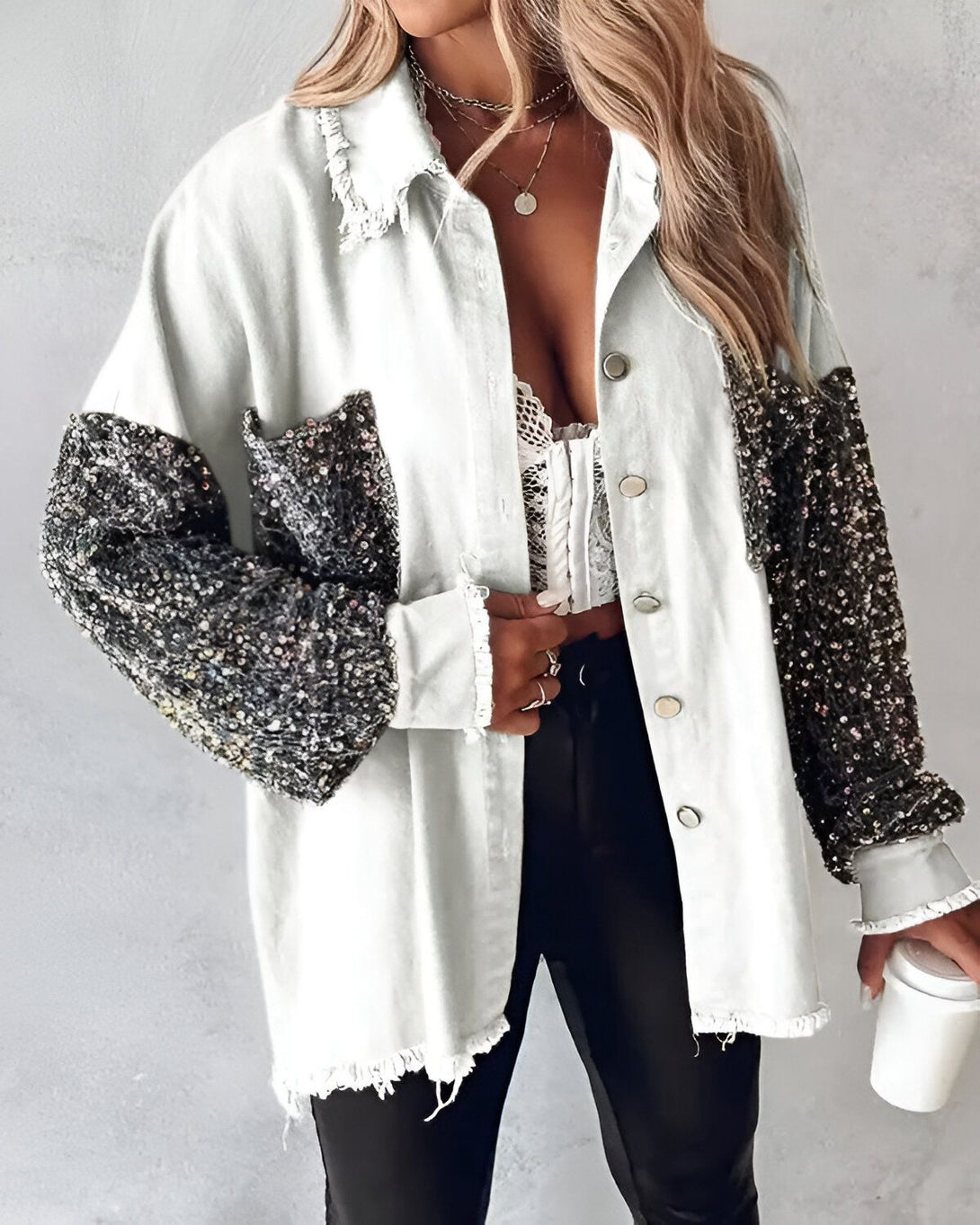 Une femme porte une veste en jean blanche avec des manches à sequins noirs. Elle est ouverte sur un haut en dentelle blanche. Elle porte également un pantalon noir et des talons noirs.