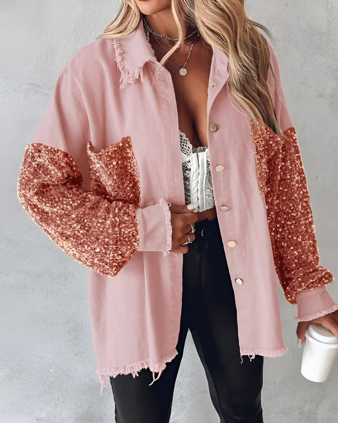 Une femme porte une chemise rose à manches longues pailletées. Elle est ouverte et on peut voir un soutien-gorge en dentelle blanche en dessous. Elle porte également un collier avec un pendentif rond et des boucles d'oreilles. Elle tient une tasse blanche à la main.