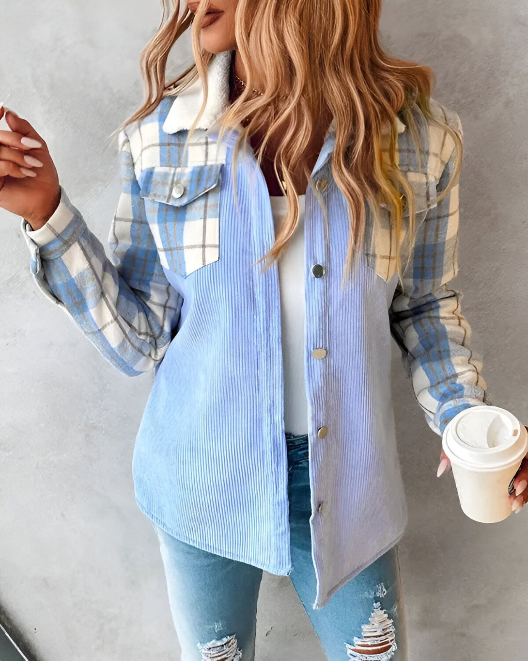 Une jeune femme porte une chemise bleue à carreaux et velours côtelé. Elle est debout et tient une tasse de café à la main.