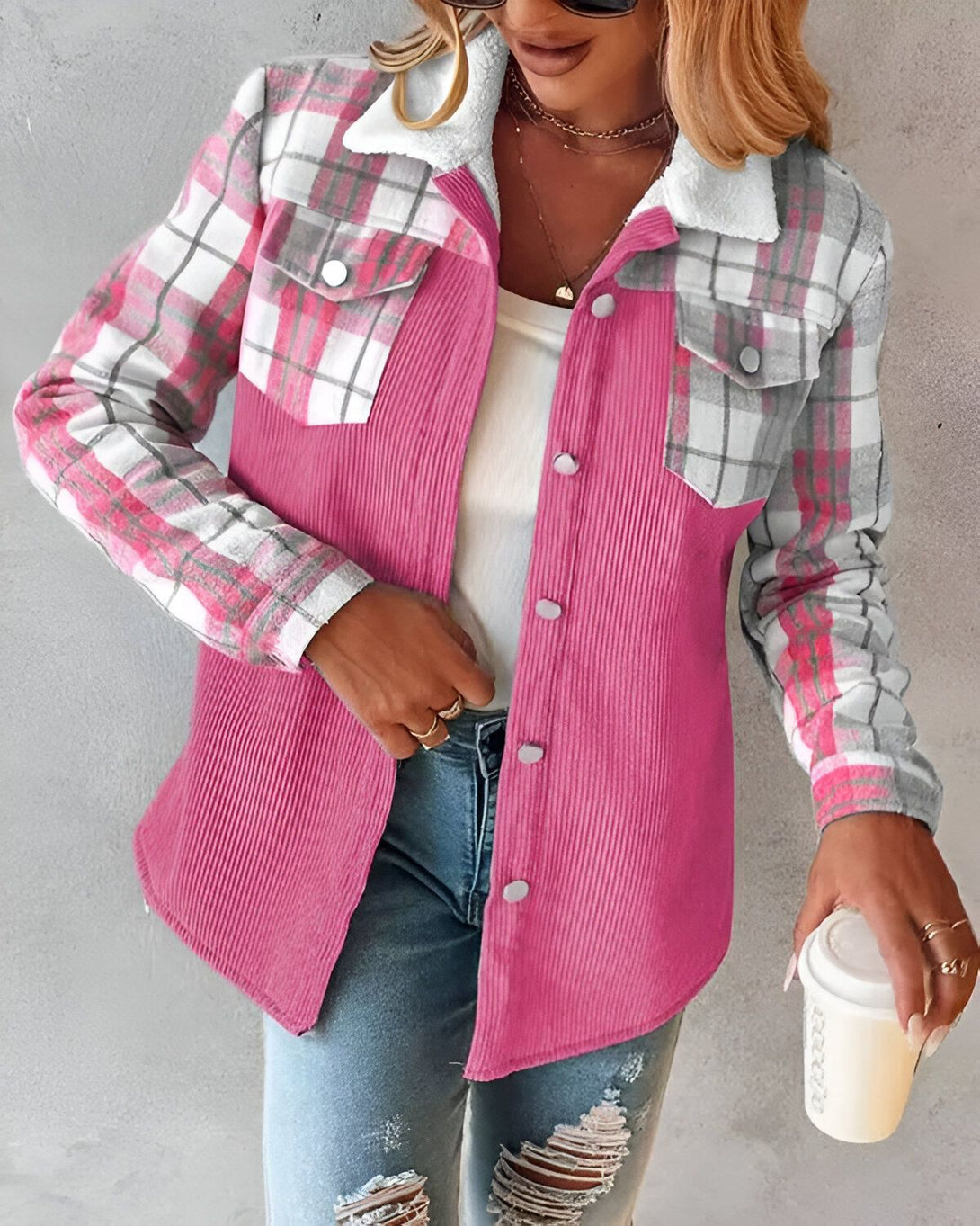Une femme porte une veste rose en velours côtelé avec des carreaux gris et blancs sur les manches et le col. Elle porte également un jean bleu et un collier.