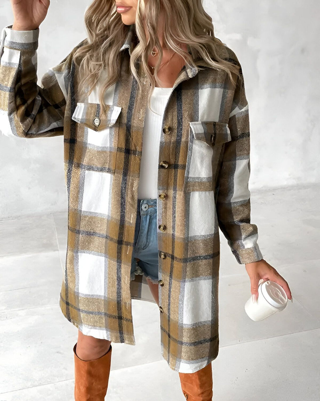 Une jeune femme porte une chemise à carreaux marron et blanche ouverte sur un t-shirt blanc. Elle porte également un short en jean bleu et des bottes marron. Elle tient une tasse blanche à la main.
