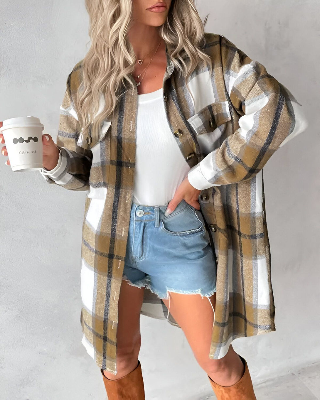 Une jeune femme porte une chemise à carreaux marron et blanche ouverte sur un débardeur blanc. Elle porte également un short en jean bleu et des bottes marron. Elle tient une tasse de café à la main.