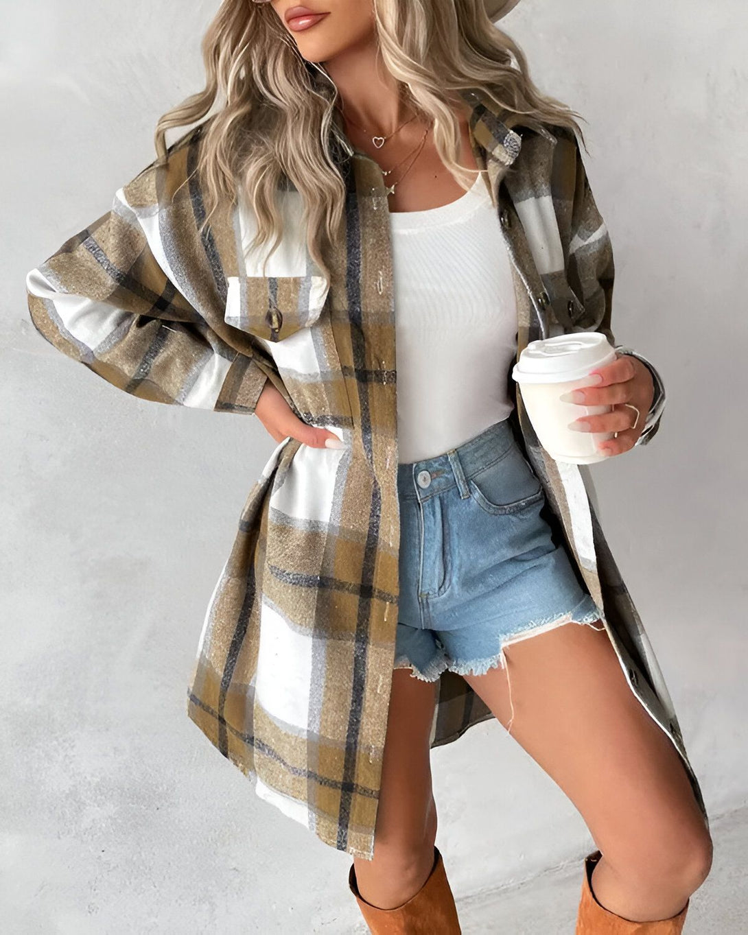 Une jeune femme porte une chemise à carreaux marron et blanche ouverte sur un débardeur blanc. Elle porte également un short en jean bleu et des bottes marron. Elle tient une tasse de café à la main.