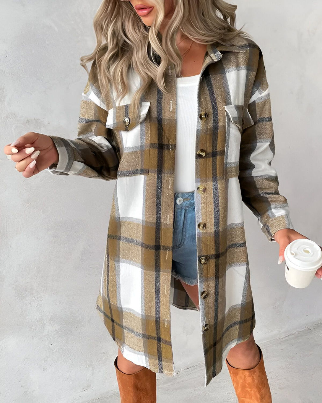 Une jeune femme porte une chemise à carreaux marron et blanche ouverte sur un t-shirt blanc. Elle porte également un jean bleu et des bottes marron. Elle tient une tasse de café à la main.