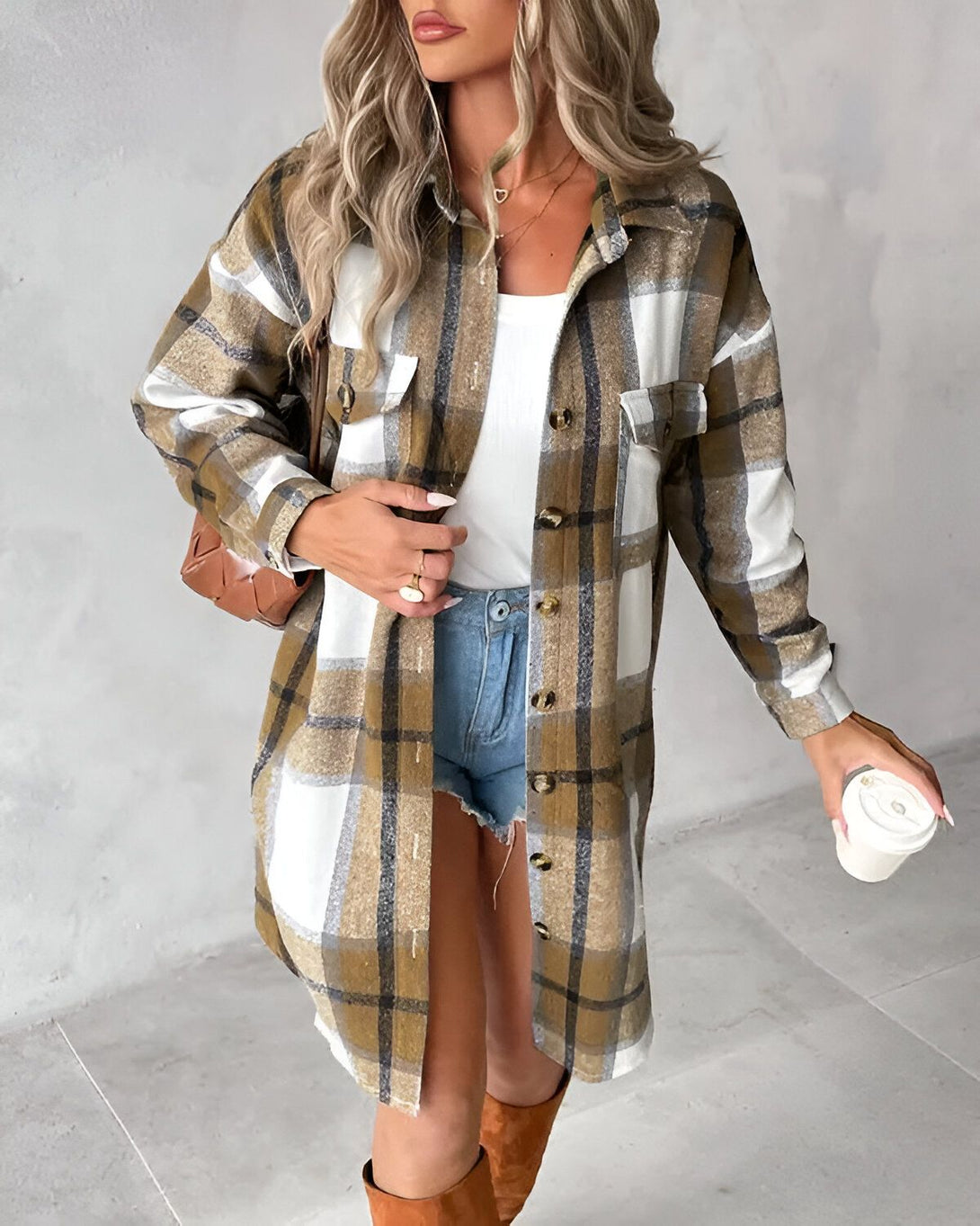 Une jeune femme porte une chemise à carreaux marron et blanche ouverte sur un débardeur blanc. Elle porte également un short en jean bleu et des bottes marron. Elle a un sac à main marron et tient une tasse de café à la main.