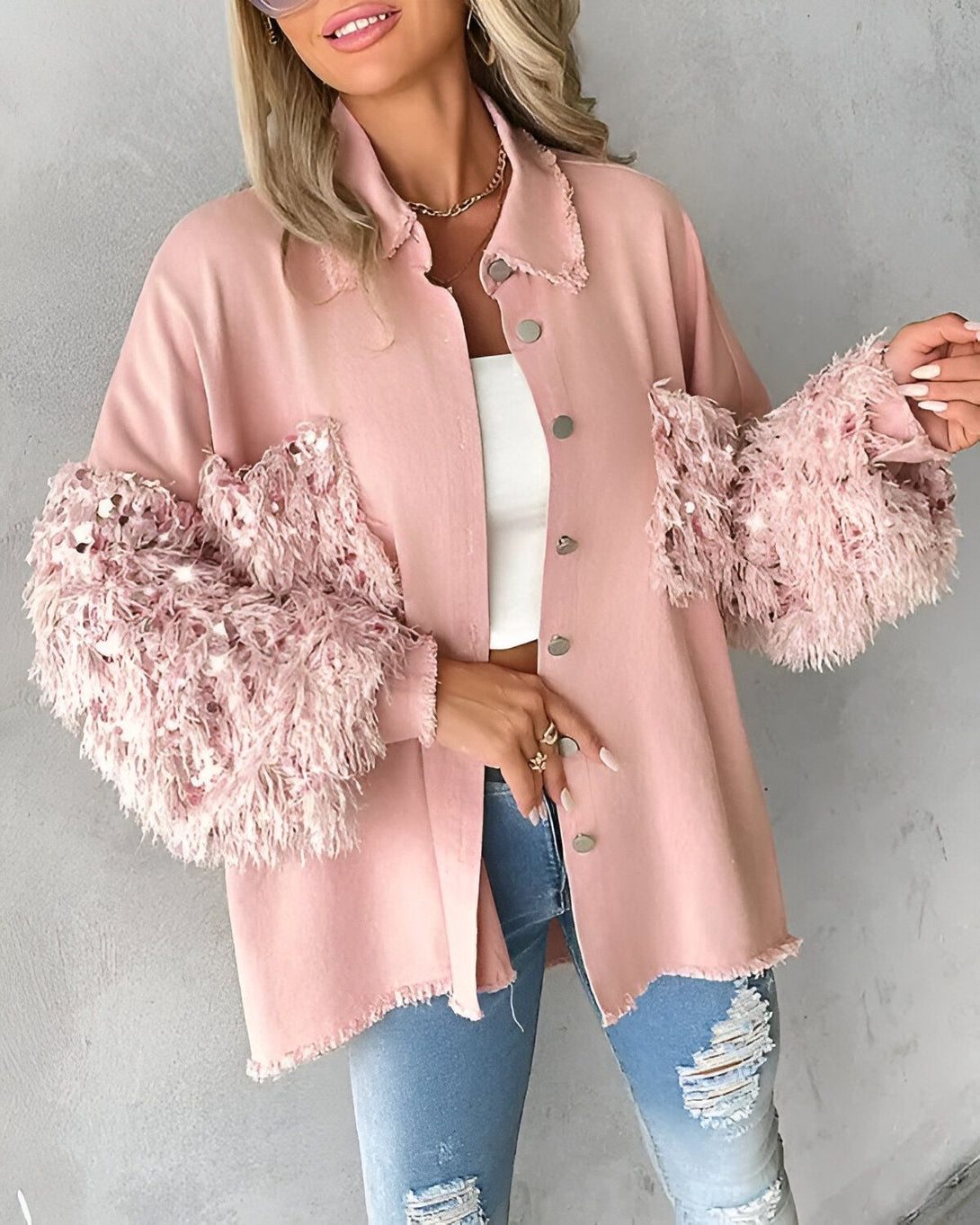 Une femme porte une veste rose à manches longues. La veste est ouverte et on peut voir qu'elle porte un débardeur blanc en dessous. Elle porte également un jean bleu déchiré et des talons hauts.