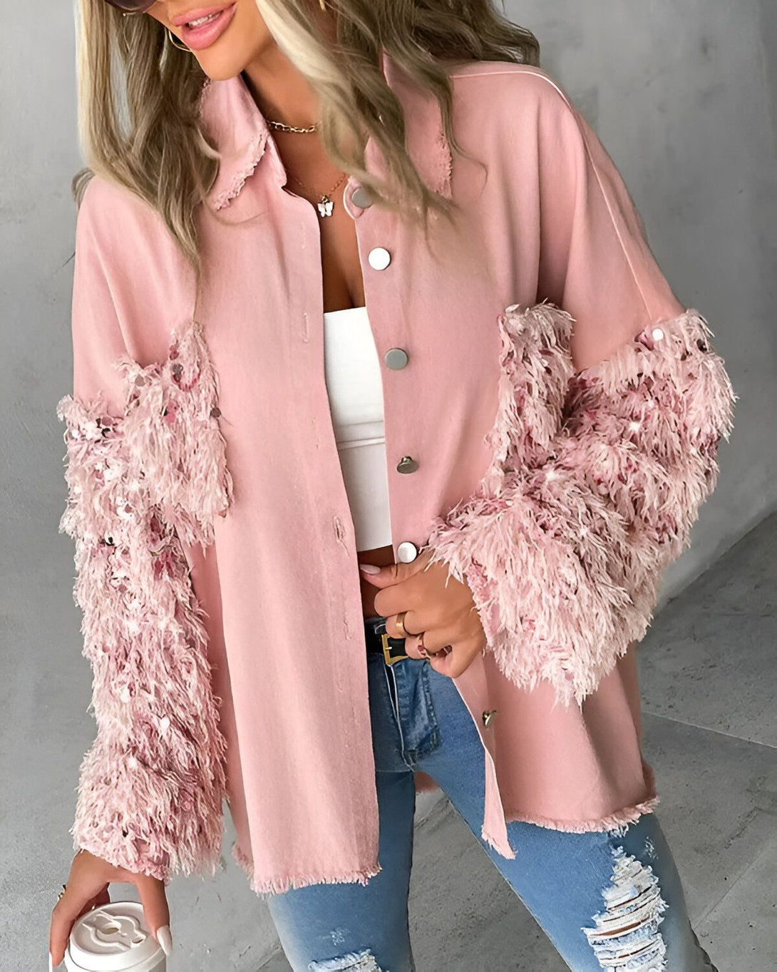 Une jeune femme porte une veste rose à manches longues. La veste est ouverte et l'on peut voir qu'elle porte un débardeur blanc en dessous. Elle porte également un jean bleu déchiré et des talons hauts. Elle a les cheveux longs et blonds et porte un maquillage léger. Elle est debout dans une rue et tient une tasse de café à la main.